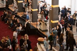 Der volle Lichthof des Gropius Bau, bei der Eröffnung der Ausstellung Ligia Lewis: Ligia Lewis: I’M NOT HERE FORRRRR… In der MItte der Besuchenden sitzt eine performenen Person auf einem Sockel.
