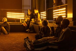 Auf dem mit rotem Teppich ausgelegten Boden des Lichthofs  des Gropius Bau sitzen Besucher*innen. Hinter ihnen ist eine Installation mit Lichtröhren.