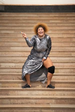 Eine weiblich gelesene Person in einem silbernen Kleid, steht auf den Treppenstufen des Foyes im Gropius Bau und singt.