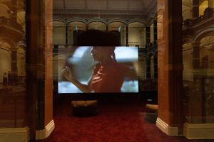 In einem Fenster, mit Blick auf den historischen Lichthof des Gropius Bau, spiegelt sich ein Bildschirm. Zu sehen ist eine Szene aus der Arbeit Arbeit „A Plot, A Scandal“ der der Künstlerin Ligia Lewis. Das angeschnittene Profil von Ligia Lewis in Bewegung ist schemenhaft zu erkennen.