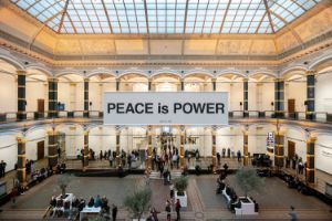 Frontalansicht des Lichthofs des Gropius Bau: In der Mitte hängte ein weißes Banner mit dem Schriftzug „Peace is Power“ in schwarzen Lettern.