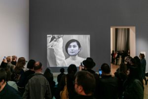A video of a young Yoko Ono is projected onto a wall in an exhibition space, with numerous people watching the video.