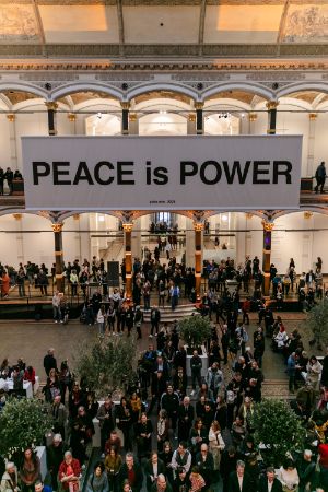 People gathered in the atrium of the Gropius Bau. Above their heads, a white banner with the words “PEACE is POWER” in black letters.