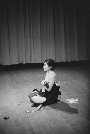 Black-and-white photograph of Yoko Ono sitting on a stage, with a pair of scissors lying next to her.