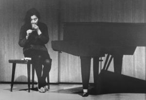 Schwarz-Weiß-Aufnahme von Yoko Ono an einem Flügel sitzend, wie sie ein Streichholz in der Hand hält.