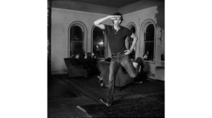 A black-and-white photograph of a person in a room with four windows, armchairs, and carpets, jumping into the air while holding one hand on their hip and the other on their head.