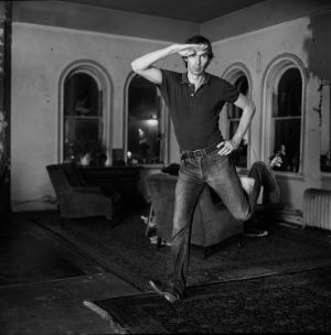 A black-and-white photograph of a person in a room with four windows, armchairs, and carpets, jumping into the air while holding one hand on their hip and the other on their head.