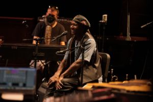 Der Musiker Hamid Drake sitzt lächelnd in einem Arrangement von Instrumenten vor einem Mikrofon. Im Hintergrund sitzt Jamie Saft am Klavier.