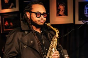 Close-up of the musician wearing black glasses and a black coat, closing his eyes and holding a saxophone in his hands.