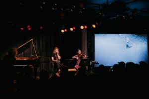 On the left Kateryna Ziabliuk on the piano, then Maryana Golovchenko on the microphone and Anna Antypova on the violin, on the right a large screen showing a large expanse of water with a raft on it.
