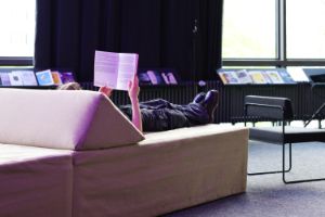 A person is lying on a modular sofa element and is reading a book. There are more books on a shelf behind the person.