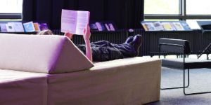 Eine Person liegt auf einem modularen Sofa-Element und liest in einem Buch. Hinter ihr sind weitere Bücher auf einem Regal zu erkennen.