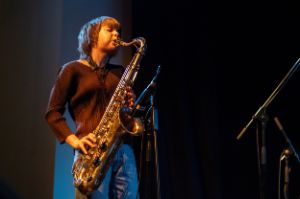 Zoh Amba on the saxophone during a live performance. There is a microphone in front of the saxophone and a black curtain in the background. Amba has her eyes closed, her cheeks puffed out as she blows into the saxophone.