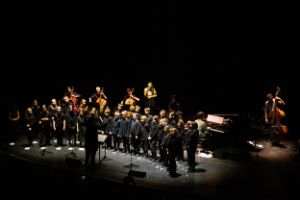In the project Apparitions, the girls' choir of the Sing-Akademie zu Berlin and the Kapellknaben of the Staats- und Domchor Berlin stand in a semi-circle on stage. In front of them in the shadow is their choirmaster. The Novembre and Bribes ensembles and three cellists can be seen in the background.