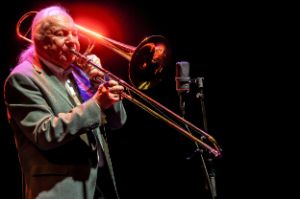 Trombonist Conny Bauer during a live performance. Bauer holds the mouthpiece to his lips, his eyes are closed. There is a microphone in front of the trombone. The musician is illuminated in red.