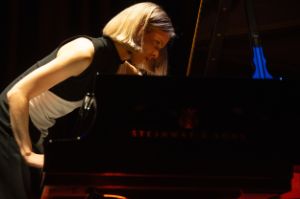 Pianist Marlies Debacker leans over a grand piano. Her gaze is directed at the piano body, her left hand seems to be reaching into the strings. The lettering Steinway & Sons can be seen on the side of the piano.