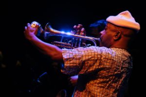 Ben LaMar Gay plays his cornet. The photo is taken from the side. LaMar Gay wears a white woollen cap. The cornet shines in the blue stage light. Individual visitors can be recognised blurred in the background.