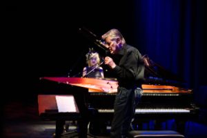 Pianist Aki Takase and pianist Alexander von Schlippenbach performing live together at the Haus der Berliner Festspiele. Alexander von Schlippenbach in the foreground, one hand on the grand piano, the other holding a microphone in front of his mouth. She sits at a second concert grand piano, which extends the first one in mirror image.
