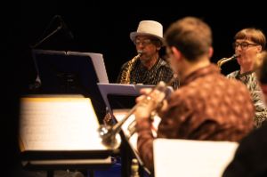 Musicians Silke Eberhard and Henry Threadgill with their ensembles Potsa Lotsa XL and Zooid during a live performance. Both musicians are seated playing their saxophones and looking at the sheet music in front of them. In the foreground, the back of another musician with a trombone can be seen in a blur.