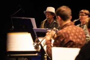 Die Musiker*innen Silke Eberhard und Henry Threadgill mit ihren Ensembles Potsa Lotsa XL und Zooid während eines Live-Auftritts. Beide Musiker*innen spielen sitzend ihr Saxofon und schauen dabei auf Noten vor ihnen. Im Vordergrund ist verschwommen der Rücken eines weiteren Musikers mit Posaune zu erkennen.