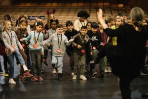 One person raises their left leg, in front of them many children try to imitate the movement.