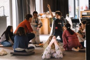 In a sitting circle, one child kneels on the floor and stretches their right index finger upwards.