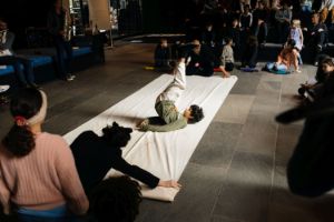 A child does a shoulder stand on a white canvas, surrounded by musicians and other children.