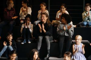 Nadin Deventer sits between children on black steps; they all applaud.