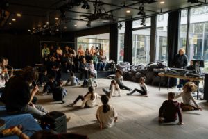 The audience sits on black steps, in front of them children are in seated movements on the floor.