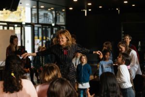 Children stand in a circle around an adult who smiles and stretches their arms out to the side.