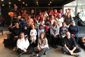 Children and adults sit in four rows on high black steps, holding red flowers in their hands.