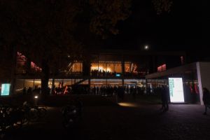 Haus der Berliner Festspiele at night. Numerous people can be seen through the windows.