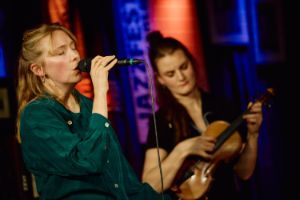 Two young musicians: one sings with her eyes closed, the other picks at the violin