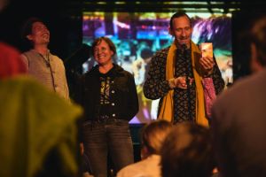 Simon Sieger, Nadin Deventer and Joel Grip stand in front of a small audience. They laugh heartily.