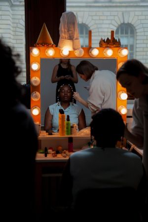 Eine Person of Colour wird geschminkt vor einem Spiegel, der mit Glühbirnen umrandet ist.