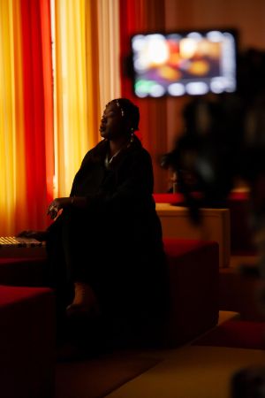 Eine Person of Colour sitzt in einem abgedunkelten Raum. Der Vorhang sperrt das Tageslicht aus.