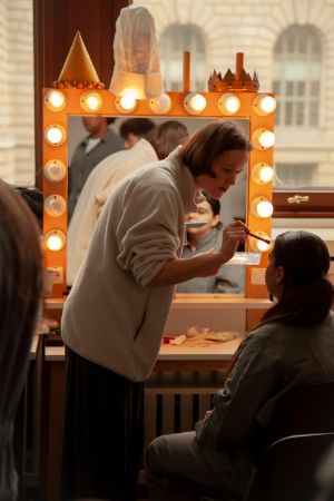 Eine Person schminkt eine andere vor einem Spiegel, der mit Glühbirnen umrahmt ist.