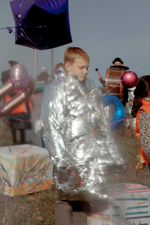Ein Kind trägt eine dicke silber glänzende Jacke. Das Kind befindet sich im Freien zwischen Musiker*inenn und Ballons.