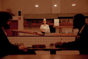 Filmszene: In einer Bar sitzen zwei Männer an einem Tisch. Am Thresen liegt ein Saiteninstrument.