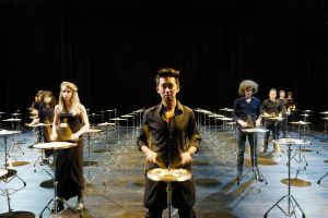 Die Mitglieder von Les Percussions de Strasbourg stehen mitten in einem Feld von 100 Becken und spielen diese.