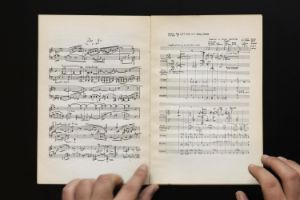 Two hands hold open a booklet with a score. At the beginning of the right-hand page is written in capital letters "Music for Left Ear in A Small Room, Number 3".
