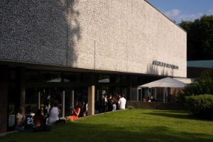 Exterior view of the Akademie der Künste building on Hanseatenweg. A few visitors are standing in front of the entrance area chatting. The sun is shining and the grass on the forecourt is green.