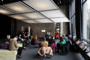 Several people sit on sofa elements in front of two speakers. To their right, a glass façade and books on deep shelves can be seen. A large light installation consisting of nine squares hangs above the visitors.