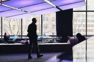 Drei Personen stehen und sitzen in einem Raum mit einer großen Glasfassade. Im Hintergrund sind Sitzelemente und tiefe Bücherregale zu erkennen. Über ihnen hängt eine große Lichtinstallation, die in hellem Lila erleuchtet und aus neun Quadraten besteht.