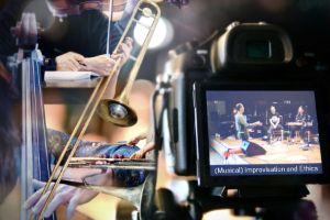 Close-up of a camera display: the recording shows four musicians on a stage talking to each other. Behind the camera is a collage of musicians making music, instruments and a person writing - the images partially overlap and merge into one another in different sizes.