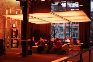 Eine leuchtende rechteckige Lichtinstallation hängt von der Decke. Darunter Sitzelemente, auf denen Menschen Platz genommen haben, und ein skulpturales Bücherregal links im Hintergrund.