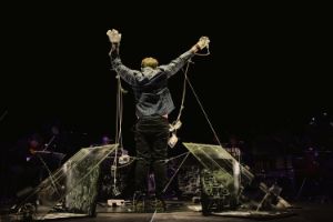 Eine Person mit verkabelten Handschuhen streckt ihre Arme nach oben. In der Luft baumeln transparente Dosen; rechts und links sind ebenfalls transparente Platten diagonal aufgestellt.