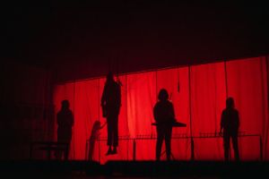Five human outlines can be recognised behind a red curtain. A human silhouette hangs in the air on several wires.