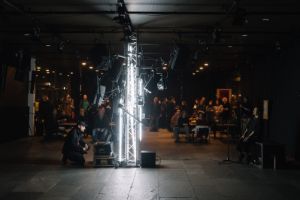 A person is sitting in front of a light installation with loudspeakers and a laptop in front of them. Another person is sitting opposite in front of a microphone. In the background is an observing audience.