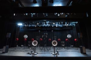 Various speaker models on a dark stage stand in spotlights.